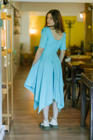 Back view of the Bubbles Dress by Cori Tailor — light blue dress with scoop neckline, puff sleeves, and handkerchief hem, worn indoors in a cozy room