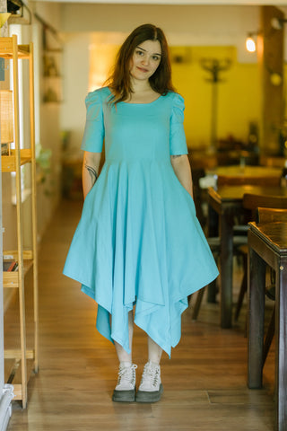 Woman wearing the Bubbles Dress by Cori Tailor — light blue dress with puff sleeves and handkerchief hem, photographed indoors in a warmly lit room