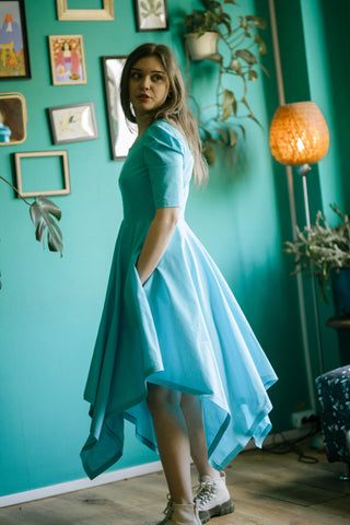 Side view of the Bubbles Dress by Cori Tailor — light blue dress with pockets, puff sleeves, and flowing handkerchief hem, worn indoors beside a teal wall and warm lamp
