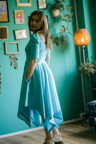 Model glancing back while wearing the Bubbles Dress by Cori Tailor — light blue dress with soft structure and pockets, shot indoors with teal wall and warm lighting