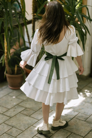 Back view of the Freya Dress by Cori Tailor, featuring puff sleeves, tiered skirt, and green bow sash, model in motion indoors