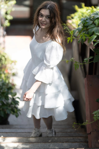 Model in the Freya Dress by Cori Tailor, smiling and standing on stairs, showcasing puff sleeves and tiered skirt in soft natural light
