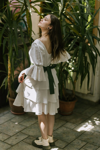 Model wearing the Freya Dress by Cori Tailor, side view showing puff sleeves, tiered skirt, and green bow sash, posed among indoor plants