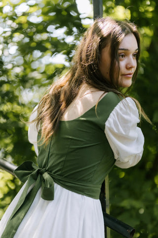 Back close-up of Heidi Dress showing green fitted bodice with tied bow and puffed white sleeves, model leaning on railing in natural setting.