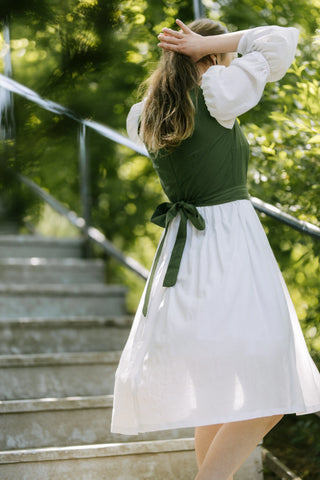 Back view of the Heidi Dress featuring green bodice with waist tie and white midi skirt, model adjusting hair on outdoor staircase.