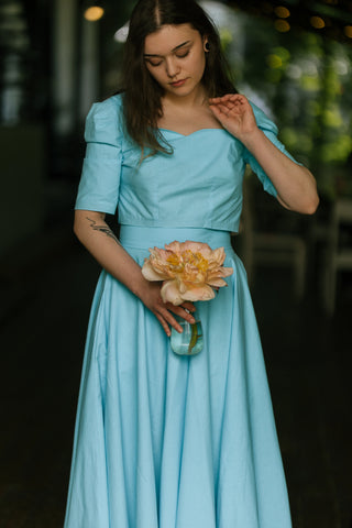 Model wearing the Jasmine Set in turquoise cotton — sweetheart neckline crop top and matching full circle skirt — holding a peach flower in a glass vase, photographed on a café terrace in Iași.