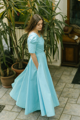 Model wearing the Jasmine Set in turquoise cotton — sweetheart neckline crop top and full circle skirt — captured in a side view while walking, indoor botanical setting.