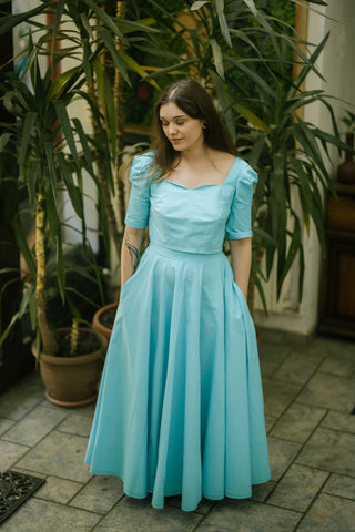 Woman wearing the Jasmine Skirt by Cori Tailor — flowing turquoise cotton skirt with a fitted waistband and full volume, styled outdoors with potted plants in the background