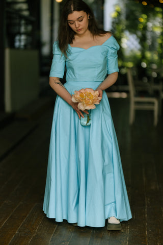 Woman wearing the Jasmine Skirt by Cori Tailor — turquoise cotton skirt styled with a fitted waistband and full volume, holding a peach flower outdoors on the terrace of Acaju cafe in Iași