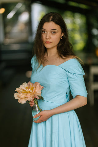 Woman wearing the Jasmine Top by Cori Tailor — turquoise cotton crop top with sweetheart neckline and puff sleeves, holding a peach flower outdoors on the terrace of Acaju cafe in Iași