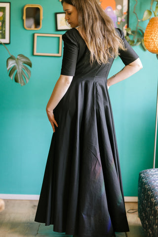 Back view of the Morticia Dress by Cori Tailor — black full-length dress with fitted bodice, sweetheart neckline, and flowing skirt, shown indoors against a green wall.