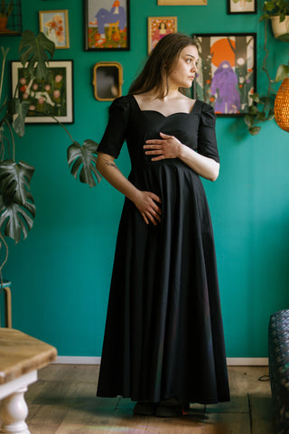 Model wearing the Morticia Dress by Cori Tailor — black full-length dress with sweetheart neckline and half sleeves, photographed indoors against a green wall with framed art.
