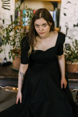 Model seated in the Morticia Dress by Cori Tailor — black sweetheart neckline dress with elbow-length sleeves, captured in a soft indoor setting with plants and books.