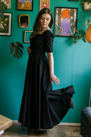 Model in the Morticia Dress by Cori Tailor — black sweetheart neckline dress with elbow-length sleeves, captured in a side pose with the skirt lifted gracefully.