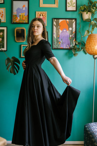Model in the Morticia Dress by Cori Tailor — black sweetheart neckline dress with elbow-length sleeves, posing in side view while holding out the flowing skirt.