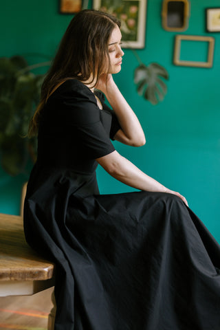 Model wearing the Morticia Dress by Cori Tailor — black sweetheart neckline dress with elbow-length sleeves, sitting on a wooden table in a serene side profile pose.