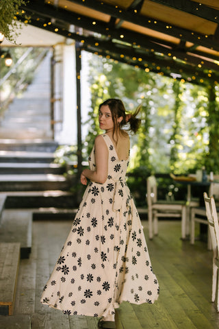 Model twirling in the Penny Lane Dress — sleeveless full-length dress with black floral print on soft beige fabric, shown from the back with tie detail, photographed on outdoor café terrace.