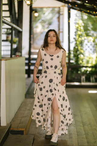Model walking toward the camera wearing the Penny Lane Dress — sleeveless floral-print maxi dress with a front slit, photographed on the Acaju terrace.
