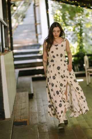  Model in motion wearing the Penny Lane Dress — beige sleeveless dress with bold black floral print and front slit, photographed walking on the Acaju terrace.