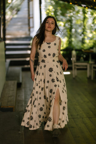Model walking in the Penny Lane Dress — sleeveless full-length beige dress with black floral print and front slit detail, photographed on outdoor café terrace.