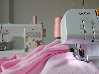 Close-up of pink fabric being sewn on a Brother overlock machine, with a second Brother sewing machine in the background at Corina’s home studio in Iași, Romania.