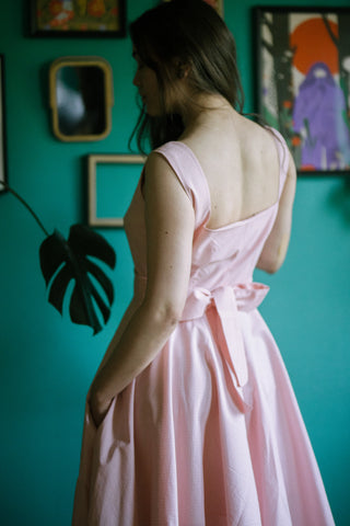 Close-up side-back view of the Sandy Dress in soft pink cotton, featuring wide straps, low back neckline, large bow detail, and full circle skirt with pockets - worn by a model against a green wall.