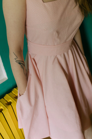Detail view of the Sandy Dress in pink cotton with small polka dots, showing the fitted bodice, waistband, full circle skirt with side pocket, and model’s arm with tattoo.