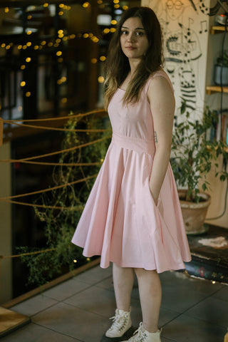 Woman wearing the Sandy Dress in soft pink cotton, featuring a square neckline, wide waistband, and full-circle skirt with pockets — captured in a softly lit indoor setting with string lights in the background.