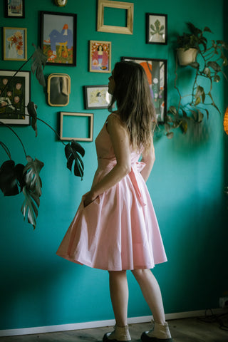 Woman wearing the Sandy Dress in soft pink cotton, shown from a side view with a wide back bow, full-circle skirt, and pockets — standing against a green wall decorated with framed art.