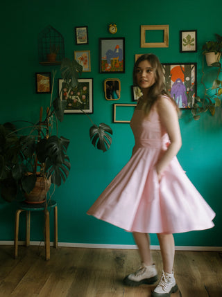 Model twirling in the Sandy Dress, a light pink cotton dress with a flared skirt and side pockets, captured mid-spin in a room with green wall and eclectic art decor.